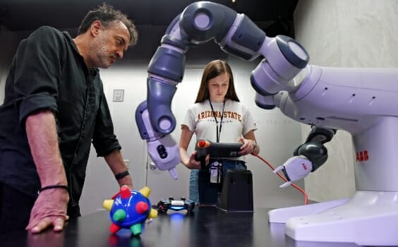 A student uses a tablet to control a robotic arm while a faculty member watches