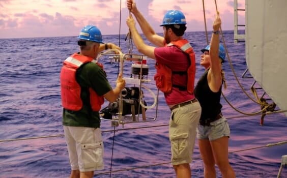 Three researchers on a boat prepare to lower a scientific instrument into the ocean