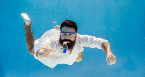 Charlie Rolsky swims underwater wearing a lab coat and safety googles, reaching for a plastic water bottle floating in front of him