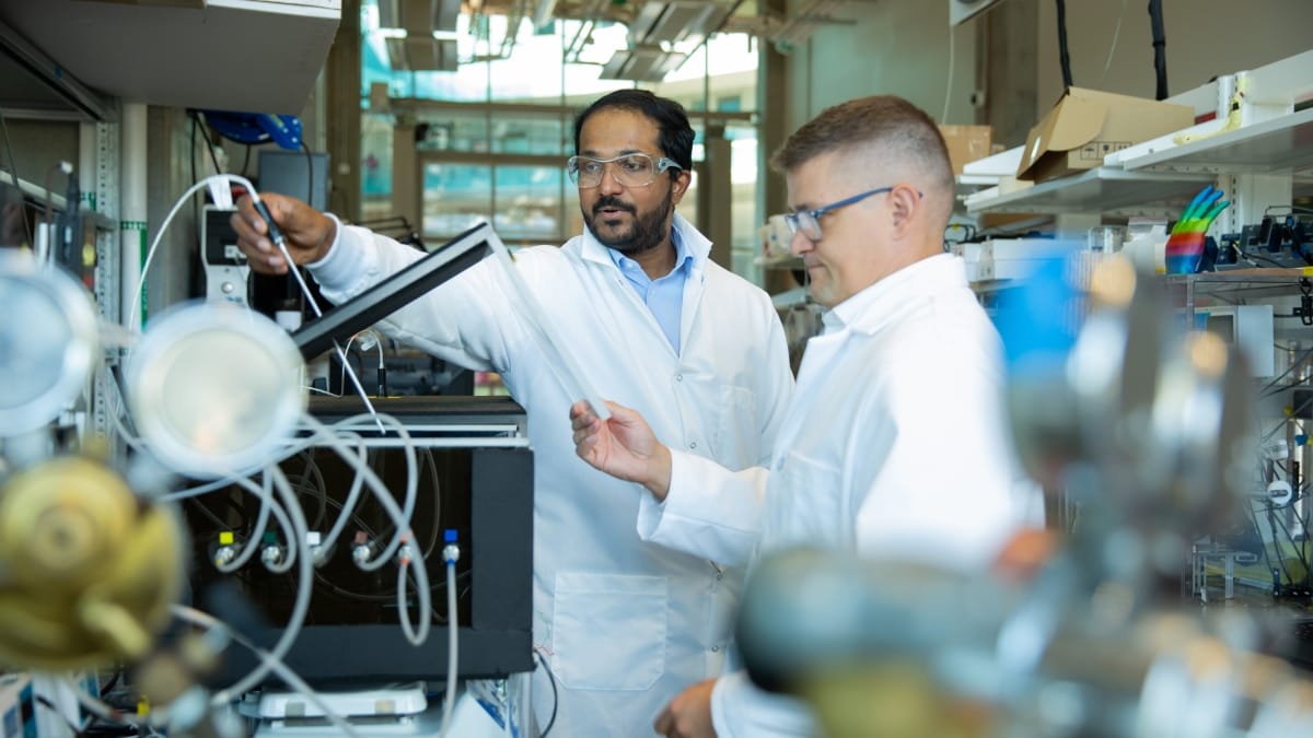 Two male researchers in lab gear examine a machine in a lab filled with technology.