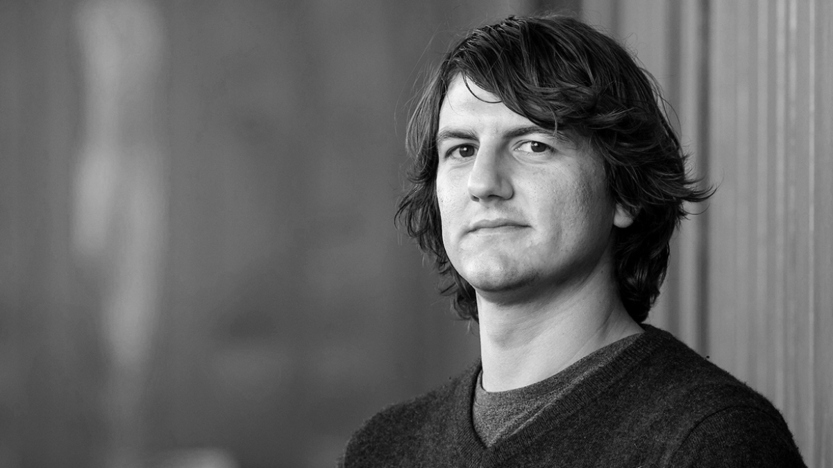 Black-and-white chest-up portrait of Adam Header with medium-length wavy hair, wearing a dark sweater against a blurred background.